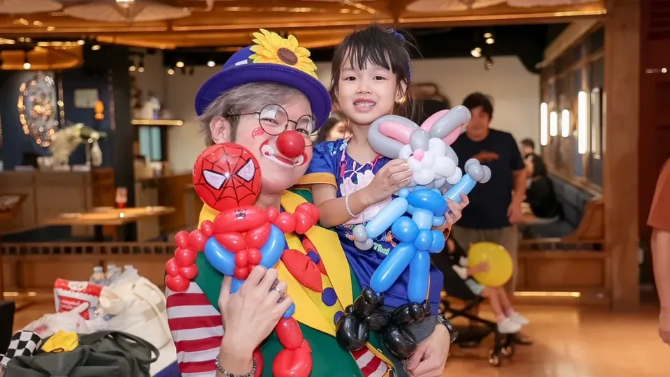 ตัวตลกถือลูกโป่งที่ปั้นเสร็จแล้วยืนคู่กับเด็กที่กำลังยิ้ม